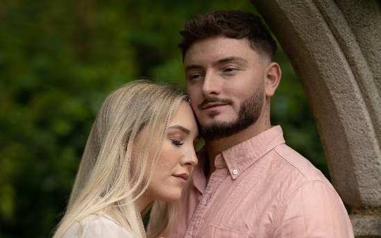 A woman with long blonde hair rests her head on the chest of a man with short brown hair and a trimmed beard. They stand close together outdoors beside a stone archway, both looking peaceful. Copyright Ashley Kew Photography - All Rights Reserved