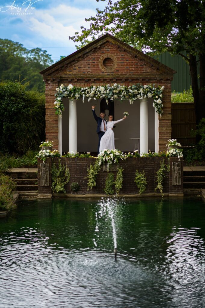 A newlywed couple poses with raised arms on the steps of a small, flower-decorated brick pavilion by a pond with a fountain, surrounded by greenery on a bright day. Copyright Ashley Kew Photography - All Rights Reserved