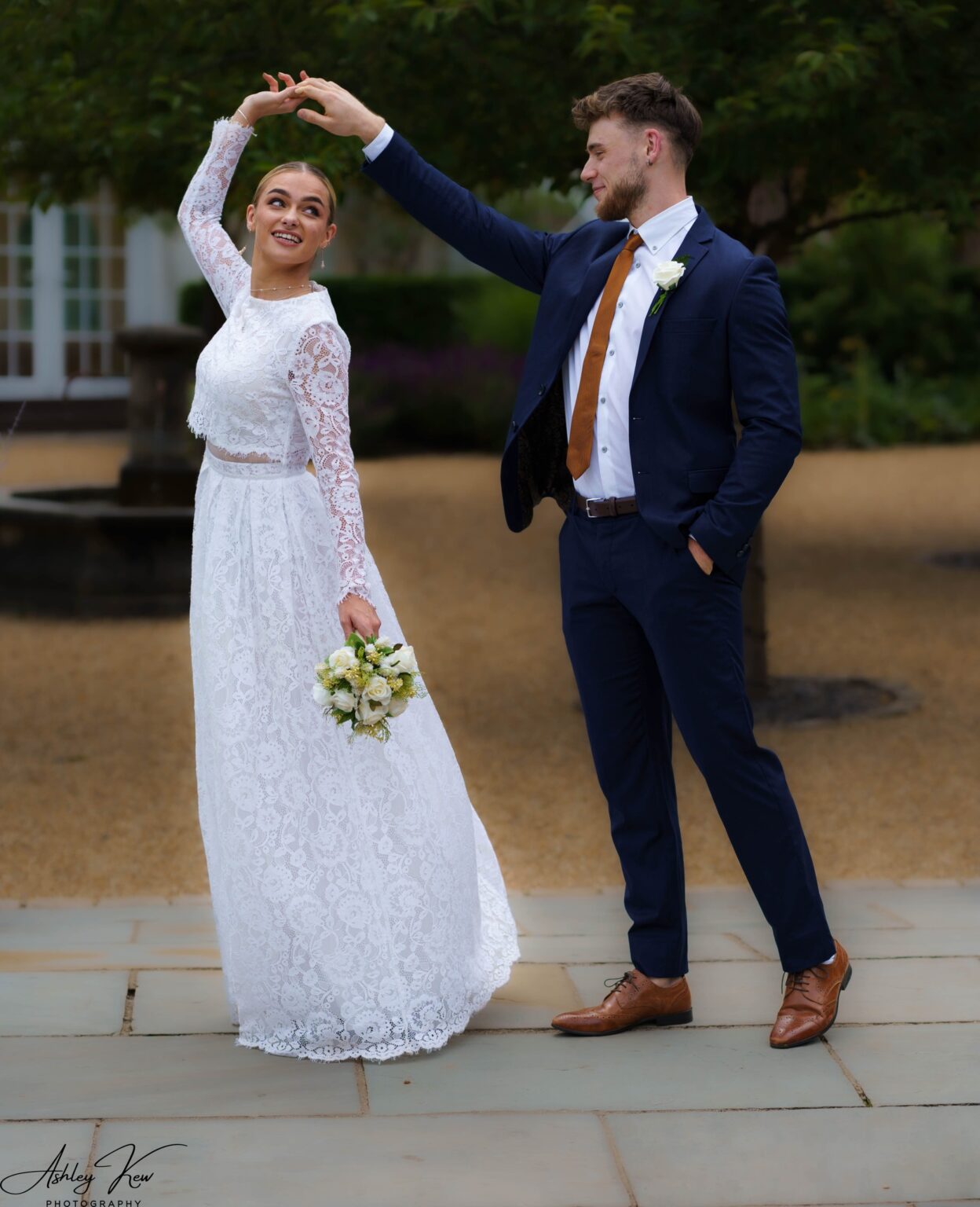 A bride in a white lace dress holds a bouquet and smiles as a groom in a navy suit and brown shoes twirls her outdoors on a stone path, with greenery in the background. Copyright Ashley Kew Photography - All Rights Reserved