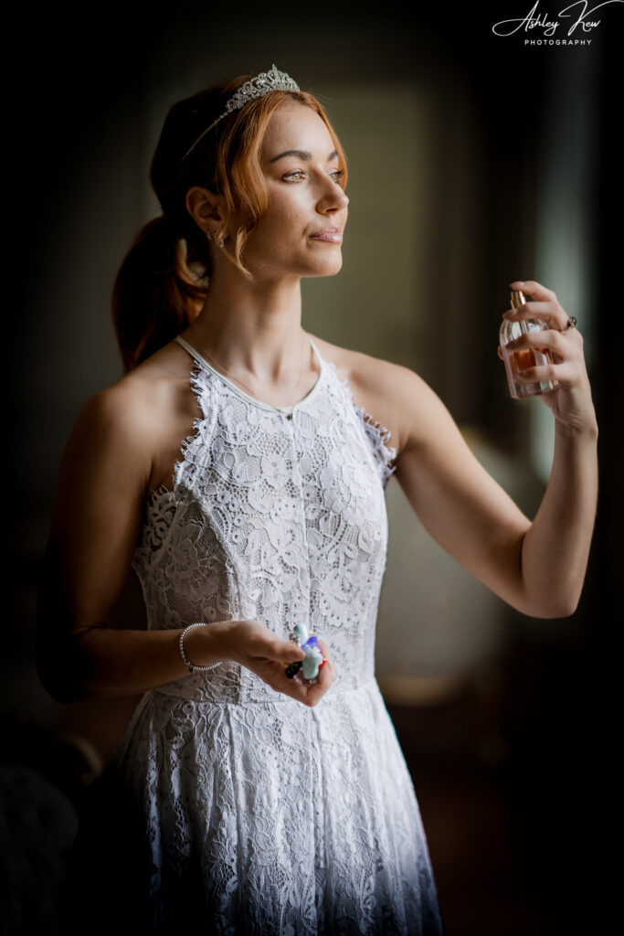 A woman in a white lace dress and tiara holds a perfume bottle, gently spraying fragrance on herself, standing in soft natural light. Copyright Ashley Kew Photography - All Rights Reserved