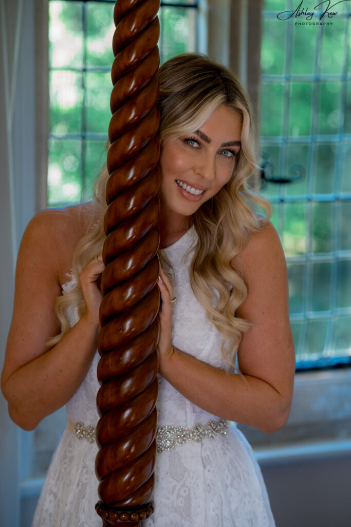 A smiling woman with long blonde hair in a white lace dress stands indoors, holding a carved wooden post. Sunlight filters through large windows with greenery visible outside. Copyright Ashley Kew Photography - All Rights Reserved