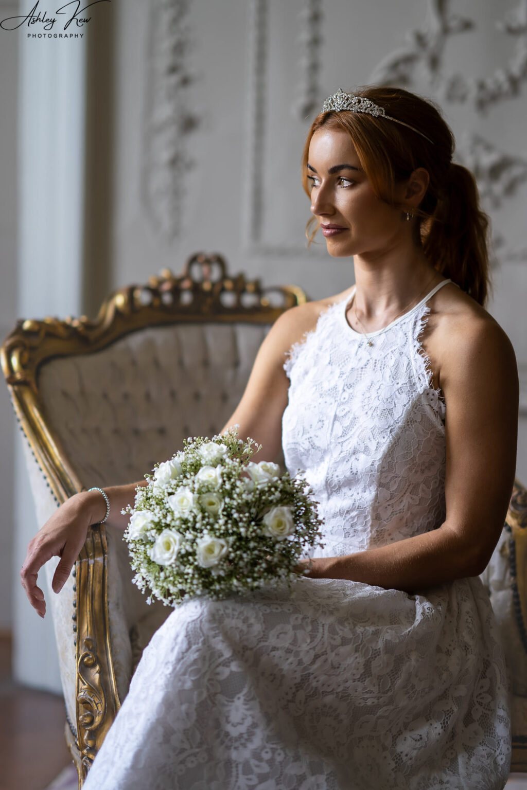 A bride in a white lace dress and tiara sits on an ornate chair, holding a bouquet of white flowers, looking thoughtfully to the side in a softly lit room. Copyright Ashley Kew Photography - All Rights Reserved