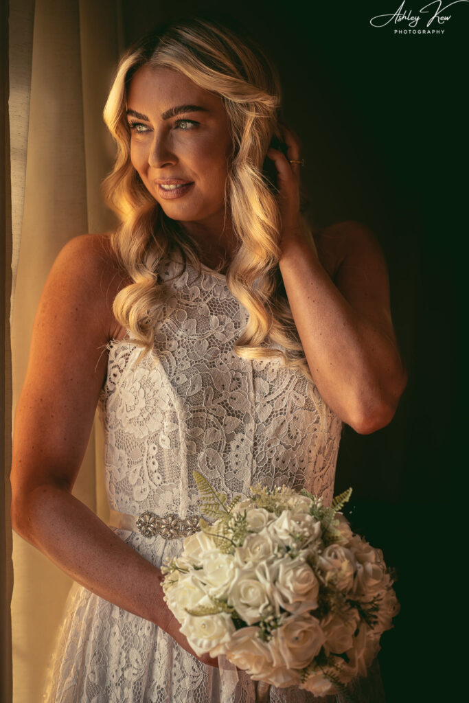 A bride in a white lace dress with wavy blonde hair stands by a window, holding a bouquet of white flowers and looking outside with a gentle smile. Warm sunlight softly illuminates her face and dress. Copyright Ashley Kew Photography - All Rights Reserved