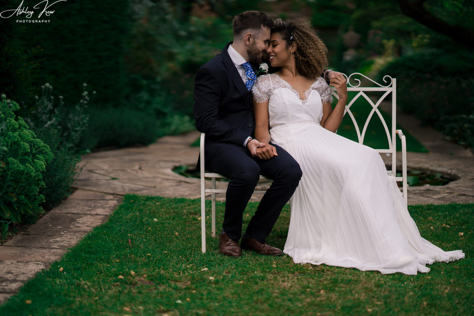 A bride and groom sit closely on a white garden bench, embracing and touching foreheads, surrounded by greenery and stone paths, dressed in wedding attire. Copyright Ashley Kew Photography - All Rights Reserved