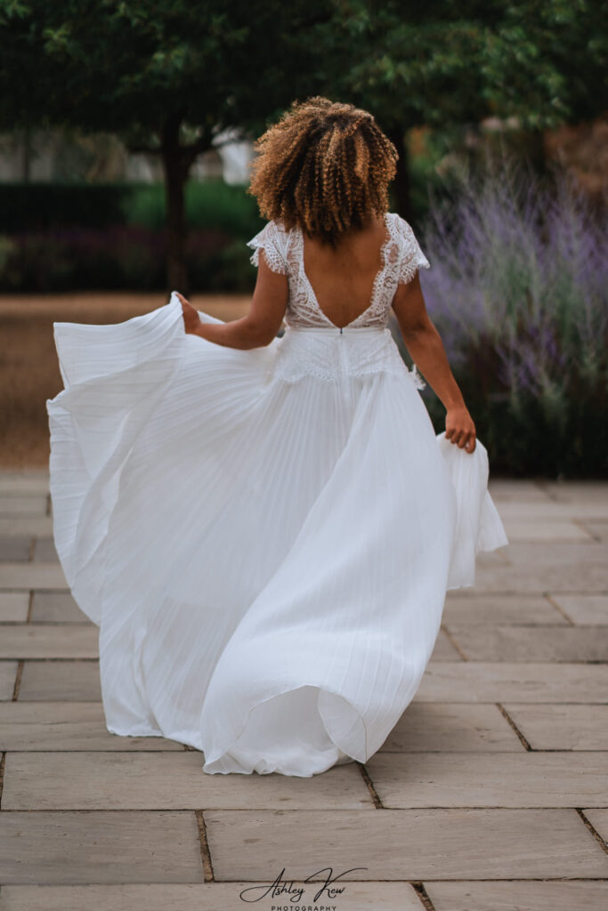 A woman with curly hair wearing a flowing white wedding dress walks outdoors on a stone path, holding her dress as it billows behind her, with green trees and purple flowers in the background. Copyright Ashley Kew Photography - All Rights Reserved