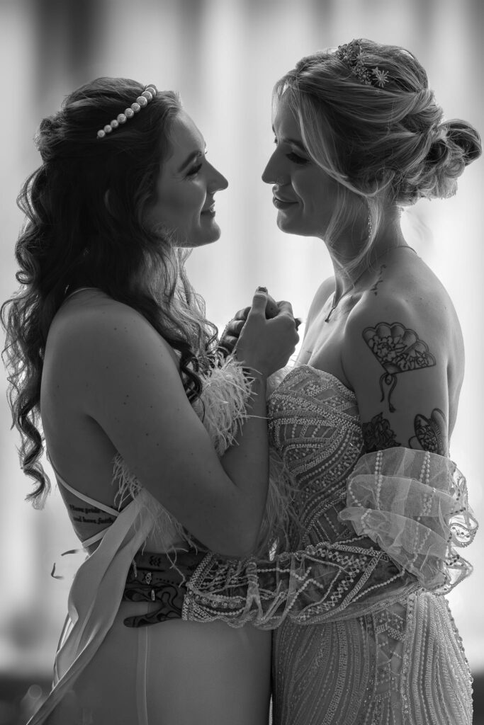 Two women in elegant dresses stand closely, gazing into each other’s eyes and smiling warmly whilst holding hands. One has loose curls with a pearl hairband; the other has an up-do, tattoos, and a jewelled tiara. Copyright Ashley Kew Photography - All Rights Reserved