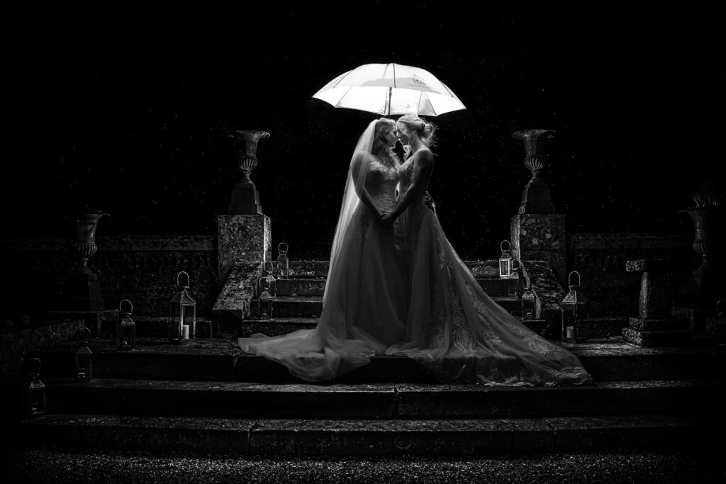Two brides in wedding dresses stand closely under a glowing umbrella at night, sharing an intimate moment in the rain on stone steps, surrounded by lanterns. The scene is dramatic and romantic in black and white. Copyright Ashley Kew Photography - All Rights Reserved