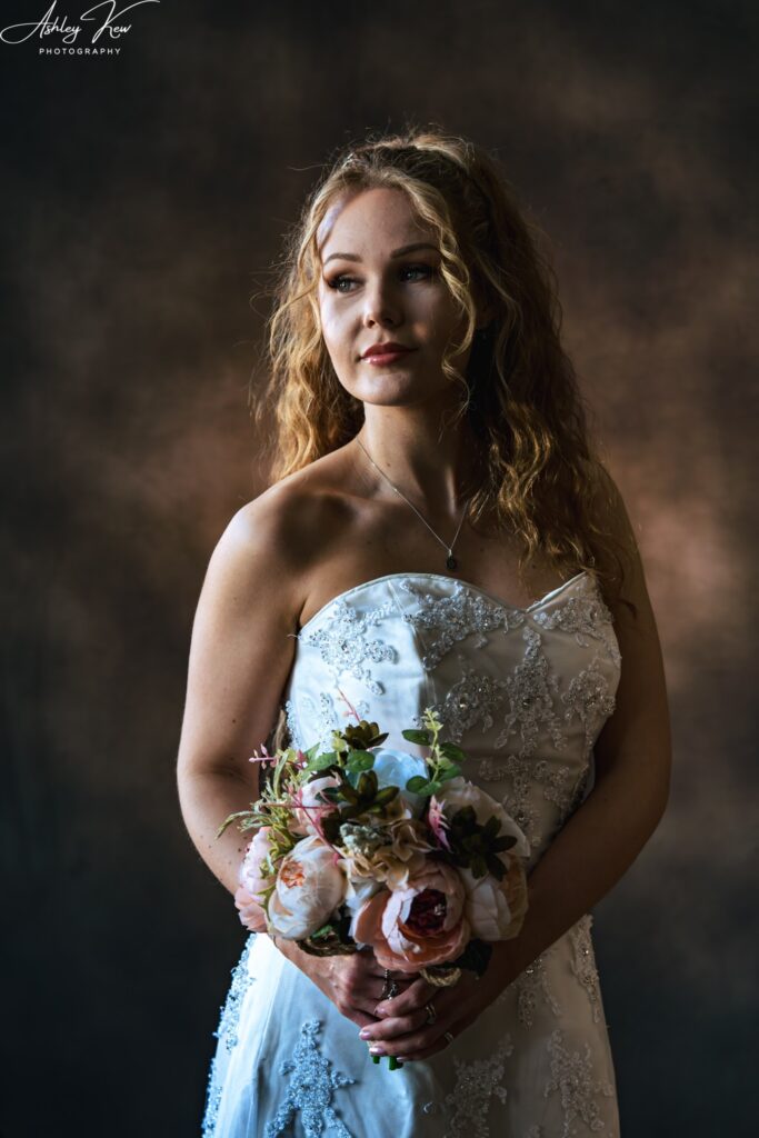 A bride in a white, strapless wedding dress holds a bouquet of pink and white flowers, gazing off to the side with a soft smile against a dark, textured background. Copyright Ashley Kew Photography - All Rights Reserved
