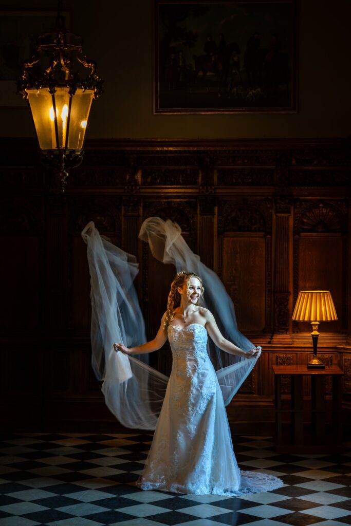 A bride in a strapless white gown poses indoors, holding her flowing veil as it catches the light. She stands on a black-and-white chequered floor, with ornate wooden panels, a glowing lamp, and a chandelier behind her. Copyright Ashley Kew Photography - All Rights Reserved