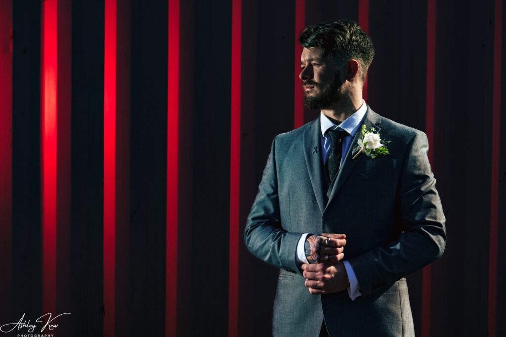 A man in a grey suit and buttonhole stands against a dark background with dramatic red vertical light beams, looking to the side with hands adjusting his cuffs. Copyright Ashley Kew Photography - All Rights Reserved
