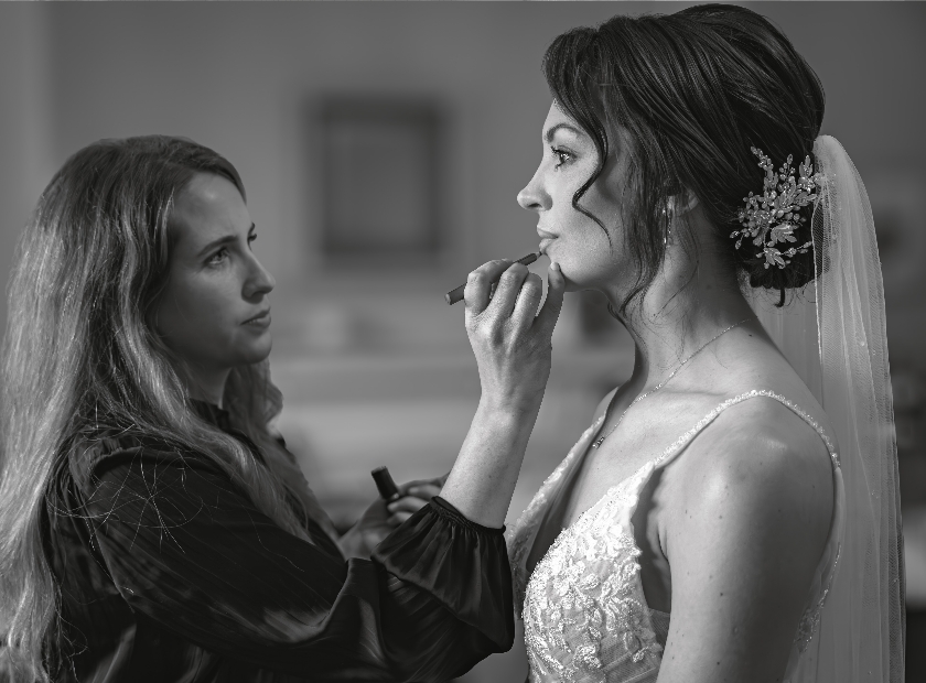A make-up artist carefully applies lipstick to a bride wearing a lace wedding dress and veil, as she prepares for her wedding. The image is in black and white. Copyright Ashley Kew Photography - All Rights Reserved