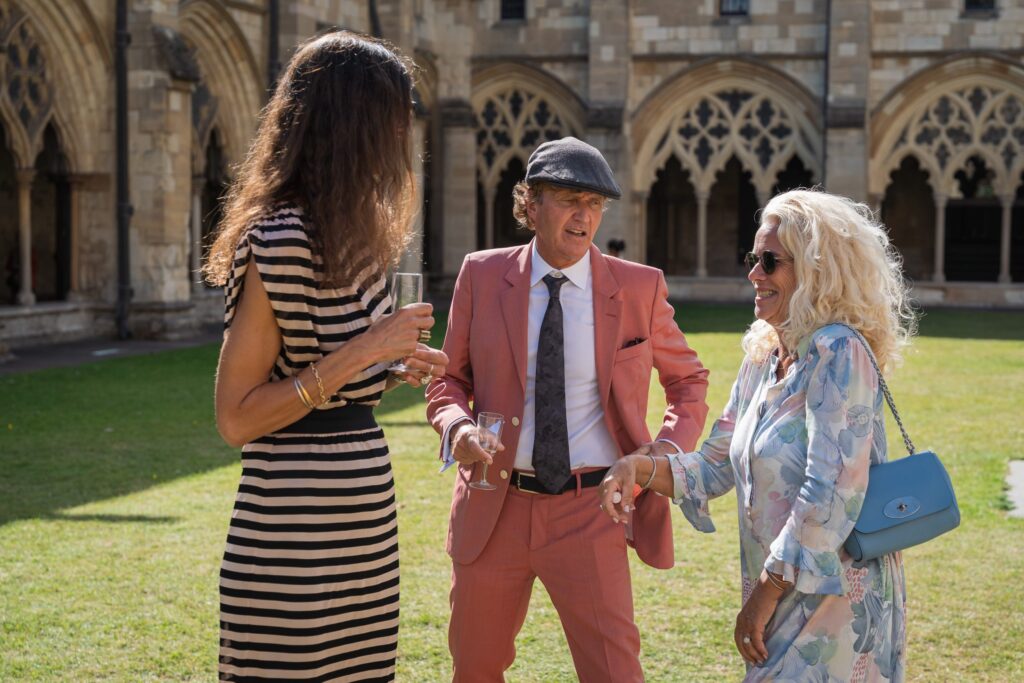 Three people are chatting outdoors near an old stone building with arched windows. One man in a pink suit and cap smiles whilst talking to two women, one in a striped dress and the other in a light floral outfit. Copyright Ashley Kew Photography - All Rights Reserved