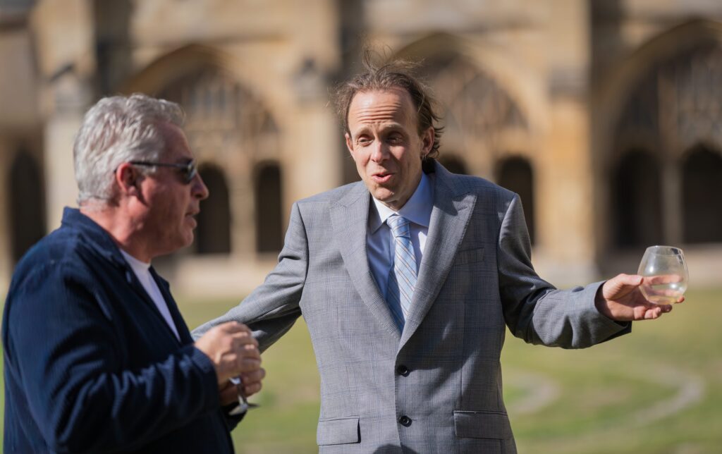 Two men stand outside talking; one in a grey suit gestures with a drink in hand, while the other in sunglasses and a dark jacket listens. They appear to be at a formal outdoor event near an historic building. Copyright Ashley Kew Photography - All Rights Reserved