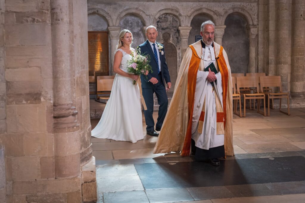 A bride holding a bouquet walks beside an older man in a suit, both smiling, as they follow a priest in a gold and white robe inside a stone church with arched walls and wooden chairs. Copyright Ashley Kew Photography - All Rights Reserved