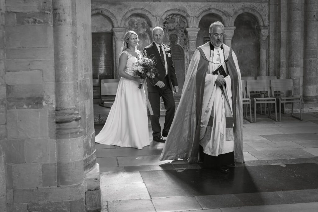A bride in a white dress, holding a bouquet, walks arm-in-arm with an older man down the aisle of a church, following a priest in ceremonial robes. The scene is in black and white. Copyright Ashley Kew Photography - All Rights Reserved