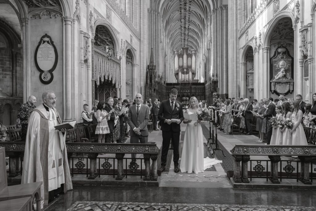 A black-and-white photo of a wedding ceremony in a grand church. The bride and groom stand at the altar with officiants and attendants whilst guests watch from pews on both sides. The ornate architecture fills the background. Copyright Ashley Kew Photography - All Rights Reserved