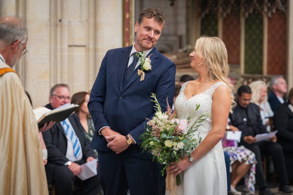 A bride and groom stand smiling at each other during a wedding ceremony, with the bride holding a bouquet of flowers. Guests are seated in the background, and an officiant stands to the left reading from a book. Copyright Ashley Kew Photography - All Rights Reserved