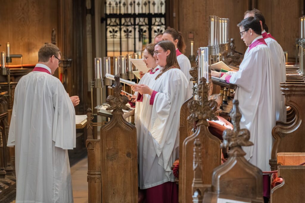 A choir dressed in white robes with red collars sings from sheet music in a wooden church choir stall, led by a conductor also in robes. The church interior features carved wood and decorative lighting. Copyright Ashley Kew Photography - All Rights Reserved