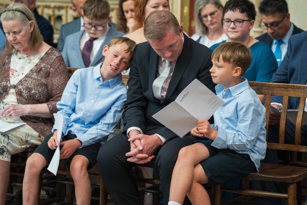 A man in a suit sits between two boys in blue shirts and shorts, looking at a programme. They are seated on a wooden bench among others at what appears to be a formal event or ceremony. Copyright Ashley Kew Photography - All Rights Reserved