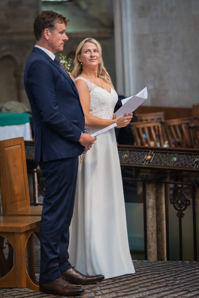 A man in a navy suit and a woman in a white wedding dress stand together indoors, holding papers and facing forwards. The woman looks up at the man, and empty wooden chairs are visible in the background. Copyright Ashley Kew Photography - All Rights Reserved