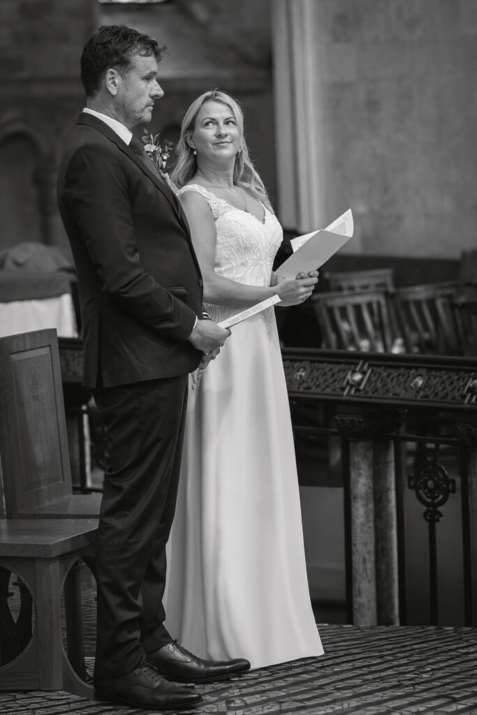 A man in a suit and a woman in a white dress stand together, holding papers, in a formal indoor setting. The woman looks up at the man, smiling. The background includes wooden chairs and ornate details. Copyright Ashley Kew Photography - All Rights Reserved