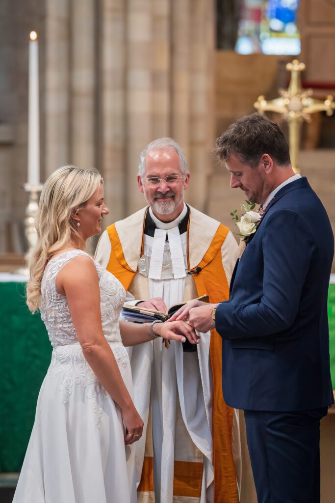 A bride and groom stand before a smiling celebrant during their wedding ceremony in a church, as the groom places a ring on the bride’s finger. Copyright Ashley Kew Photography - All Rights Reserved