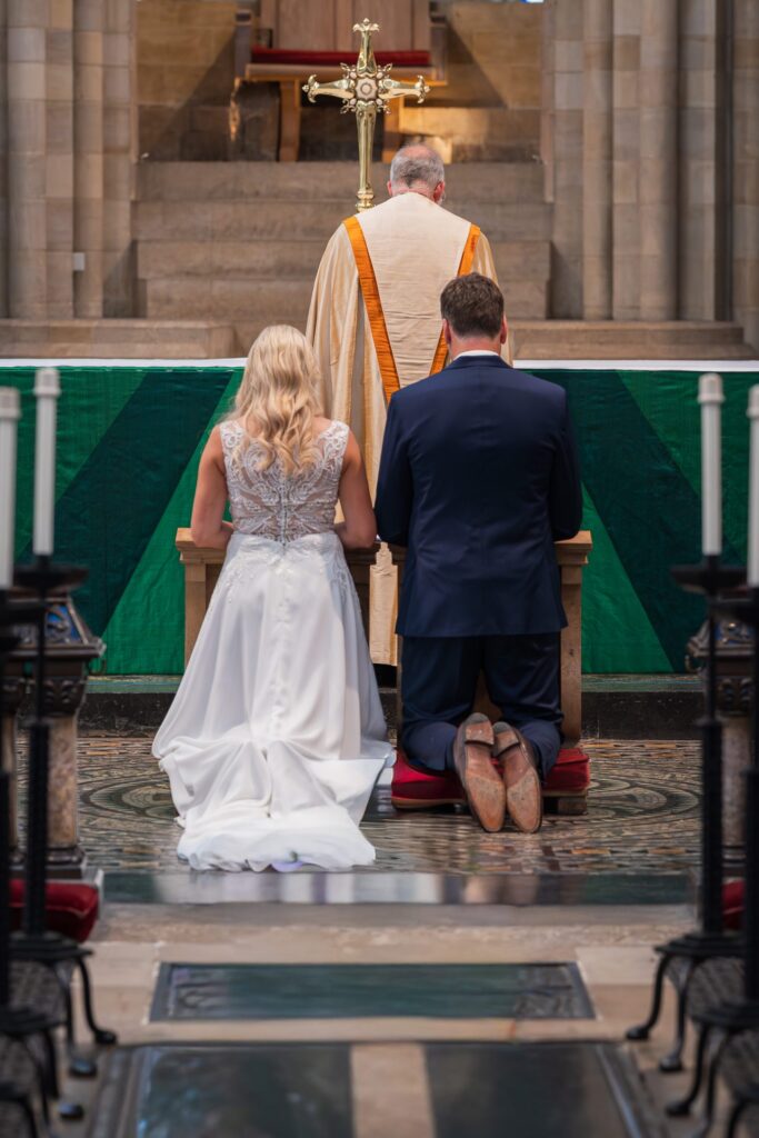 A bride and groom kneel side by side at an altar in a church, facing a priest in a white robe who stands before them. The bride wears a white gown, and the groom is in a navy blue suit. Copyright Ashley Kew Photography - All Rights Reserved