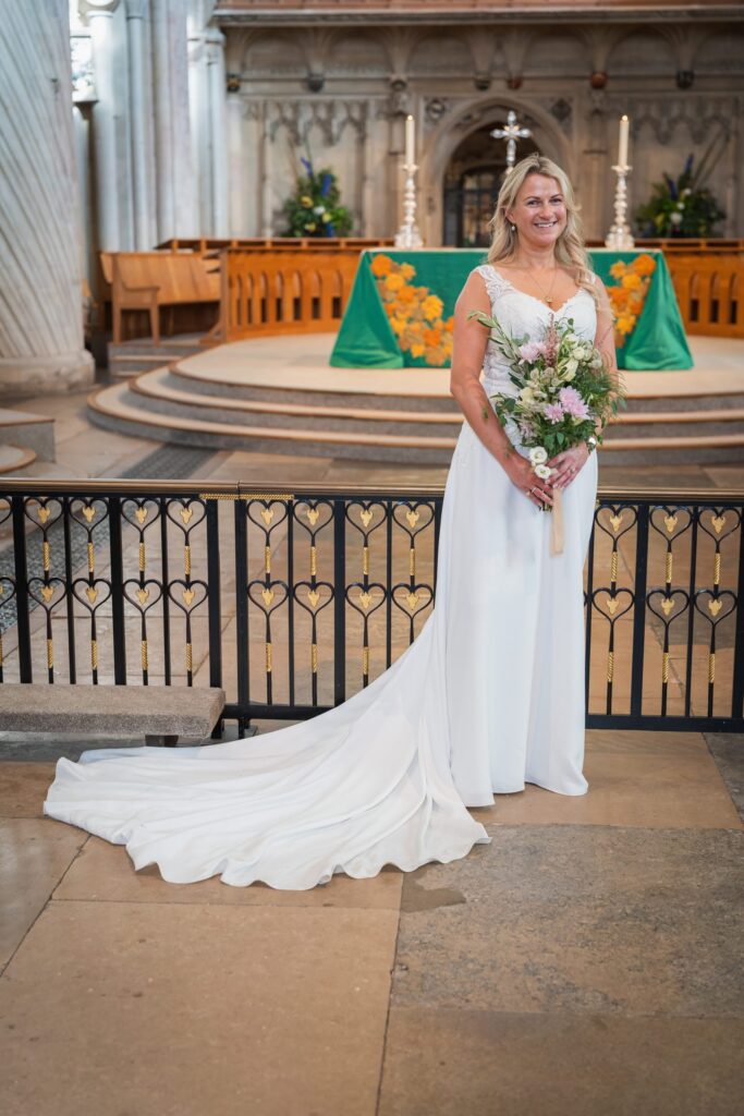 A smiling bride in a white wedding dress with a long train holds a bouquet of flowers, standing in front of an ornate church altar decorated with green and orange cloth and floral arrangements. Copyright Ashley Kew Photography - All Rights Reserved