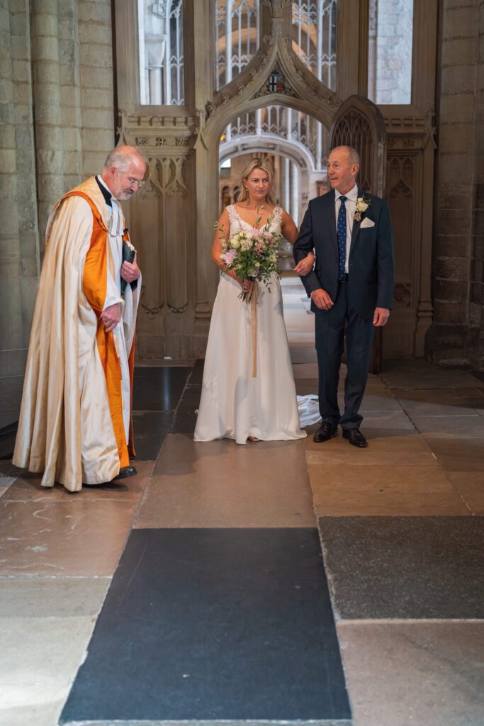 A bride in a white dress holds a bouquet and is escorted by an older man in a suit down a stone aisle inside a grand church, whilst a priest in robes stands nearby. Copyright Ashley Kew Photography - All Rights Reserved
