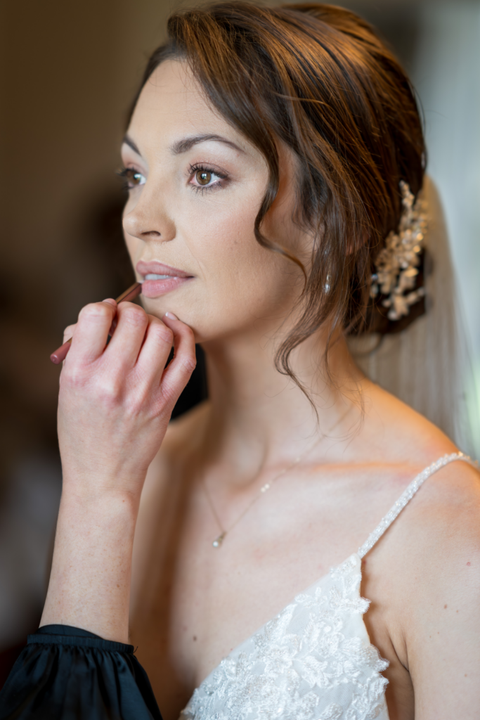 A woman in a white lace dress has her make-up done, with a make-up artist applying lipstick. She has brown hair styled with a decorative hairpiece and looks thoughtfully ahead. Copyright Ashley Kew Photography - All Rights Reserved