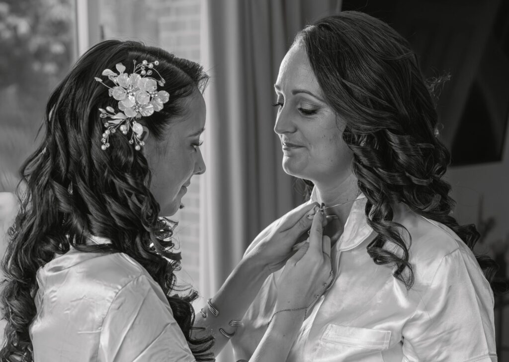 Two women with curled hair in satin dressing gowns stand close indoors. One woman helps the other put on a necklace, both smiling softly. The scene appears intimate and caring, likely on a special occasion. Copyright Ashley Kew Photography - All Rights Reserved