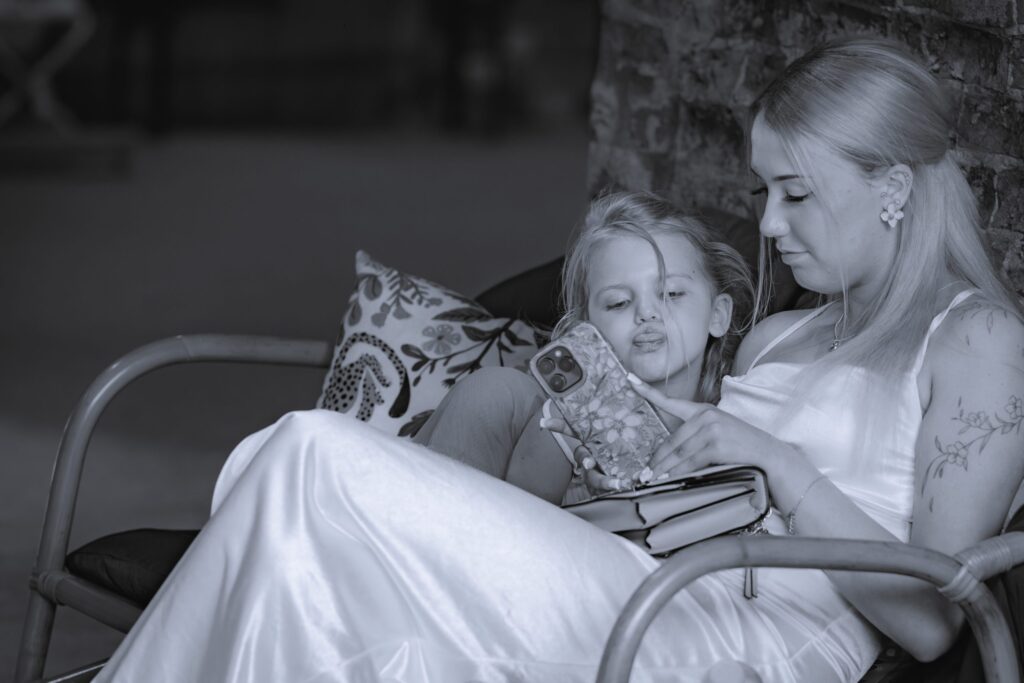 A woman in a satin dress sits on a cushioned chair next to a young girl, both looking at a mobile phone together and smiling. The scene is cosy, with a brick wall and patterned cushion in the background. Copyright Ashley Kew Photography - All Rights Reserved