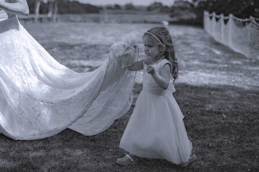 A young girl in a dress walks across grass, holding onto the train of a woman's wedding dress, outdoors on a sunny day. Copyright Ashley Kew Photography - All Rights Reserved
