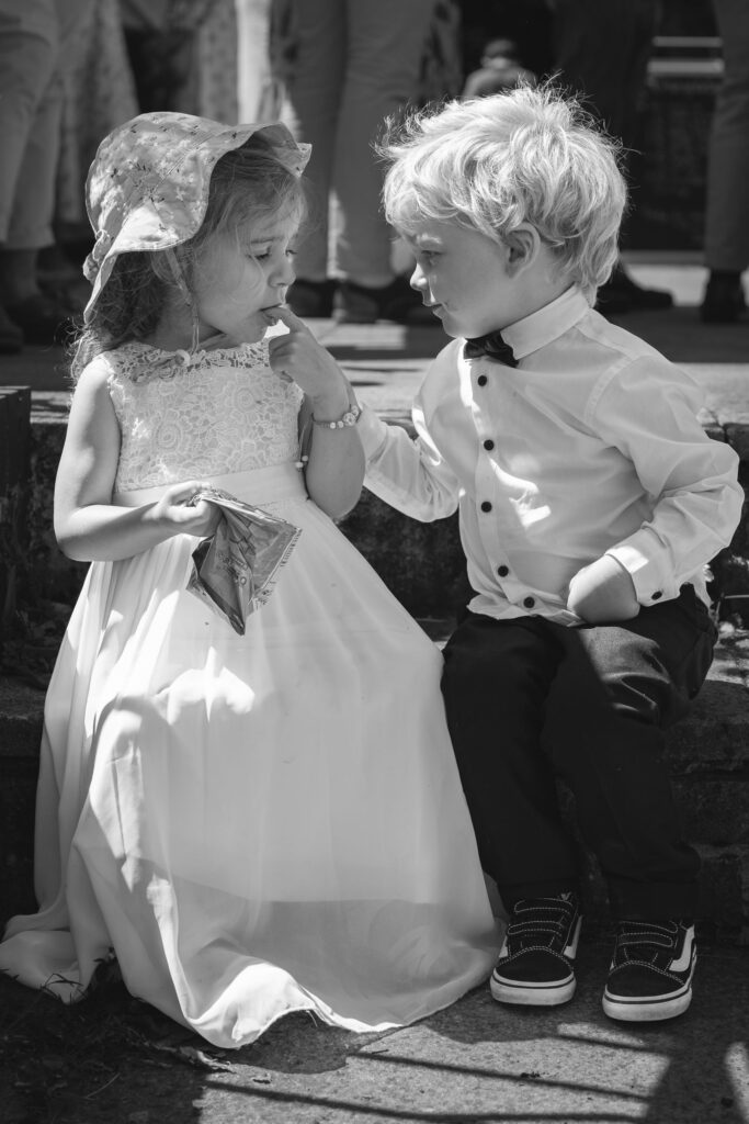 A young girl in a white dress and sunhat shares a snack with a boy in a dress shirt and bow tie as they sit together on stone steps. The background shows people standing in soft focus. Copyright Ashley Kew Photography - All Rights Reserved