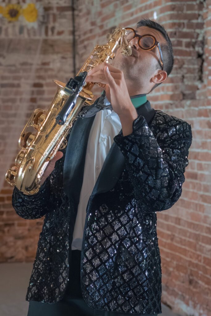 A person wearing a black sequinned jacket and large glasses plays a golden saxophone with eyes closed, standing in front of a brick wall. Copyright Ashley Kew Photography - All Rights Reserved
