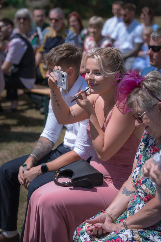 A woman in a pink dress and fascinator sits among a crowd at an outdoor event, holding a mobile to take a picture while applying lipstick with a small mirror. Other attendees sit behind her, watching. Copyright Ashley Kew Photography - All Rights Reserved