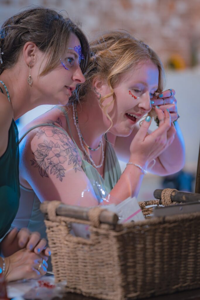Two women lean over a wicker basket, one applying face gems near her eye while the other looks on. They both wear sparkly face decorations and light dresses, focusing intently, possibly preparing for a festive event. Copyright Ashley Kew Photography - All Rights Reserved
