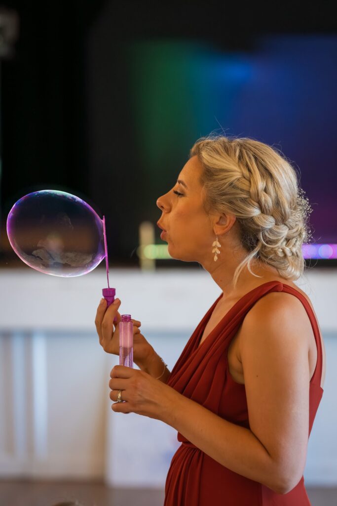 A woman in a red dress with blonde plaited hair is blowing a soap bubble indoors, holding a bubble wand and a bottle of bubble solution. The background is softly blurred with colourful lighting. Copyright Ashley Kew Photography - All Rights Reserved