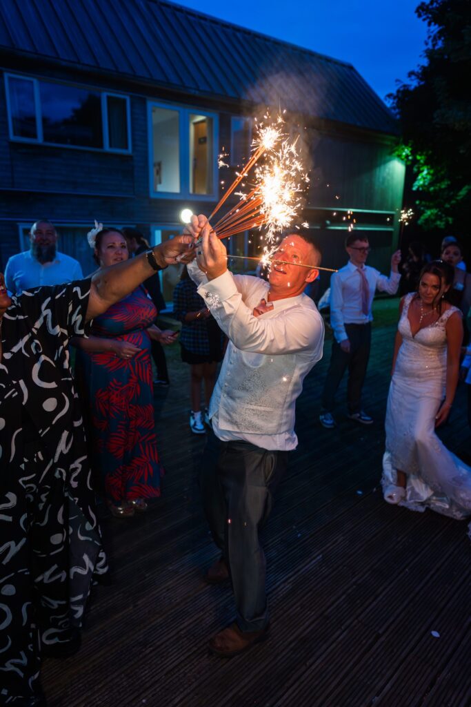 A man in a white shirt holds lit sparklers and smiles at an outdoor evening celebration, surrounded by people in festive attire, including a woman in a wedding dress. Copyright Ashley Kew Photography - All Rights Reserved