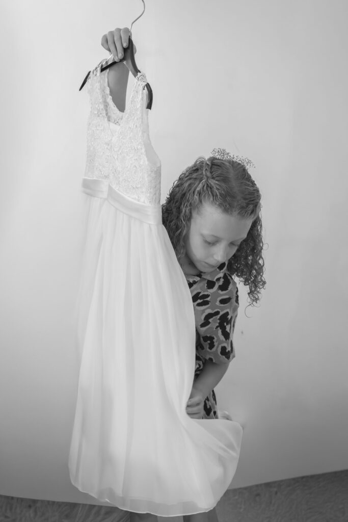 A young girl with curly hair and a leopard print shirt holds up a white sleeveless dress on a hanger, looking down at the dress. The image is in black and white. Copyright Ashley Kew Photography - All Rights Reserved