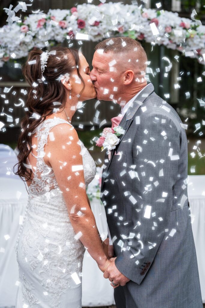 A bride and groom kiss while holding hands at their wedding, surrounded by falling white confetti. The bride wears a white lace dress; the groom is in a grey suit with a buttonhole. A floral arrangement is in the background. Copyright Ashley Kew Photography - All Rights Reserved