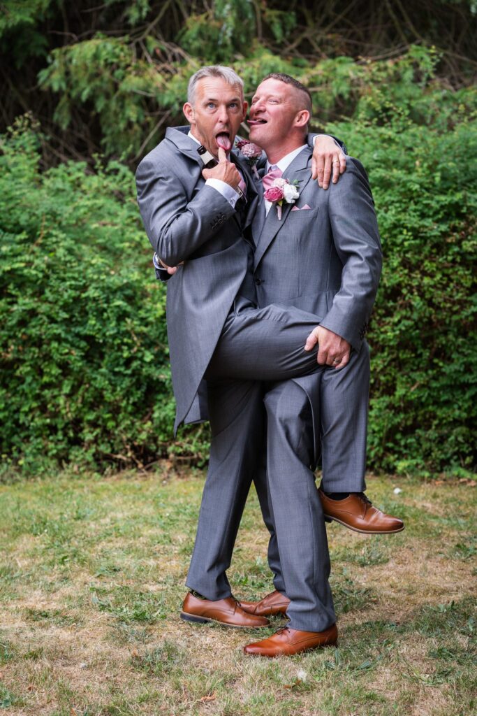 Two men in grey suits pose playfully outdoors; one pulls a funny face and sticks out his tongue while being held, leg raised, by the other man. Both wear buttonholes and brown shoes, standing on grass with greenery behind. Copyright Ashley Kew Photography - All Rights Reserved