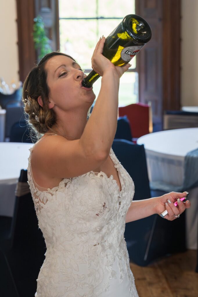 A bride in a white lace wedding dress drinks from a large green bottle while holding a lighter, standing indoors near tables and a large window. Copyright Ashley Kew Photography - All Rights Reserved