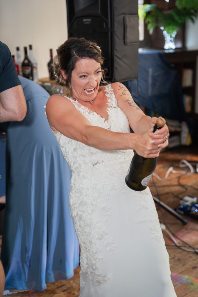 A bride in a white lace wedding dress excitedly opens a bottle of champagne indoors, laughing with wide eyes. She’s surrounded by decorations and people, creating a lively celebration atmosphere. Copyright Ashley Kew Photography - All Rights Reserved