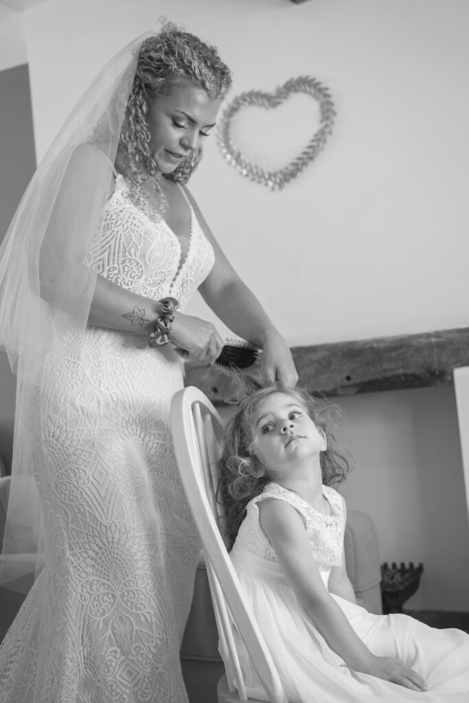 A bride in a wedding dress and veil gently brushes the hair of a young girl in a white dress, who sits on a chair. A heart-shaped decoration hangs on the wall behind them. The photo is in black and white. Copyright Ashley Kew Photography - All Rights Reserved
