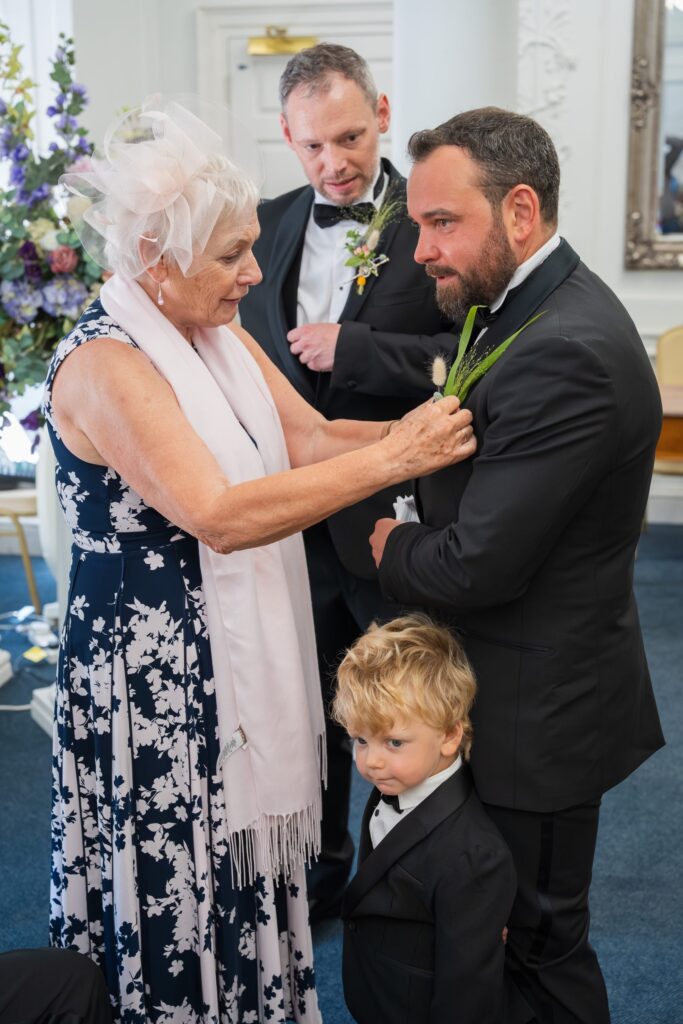 An older woman pins a buttonhole on a man’s dinner jacket while a young boy in a suit stands in front of him. Another man in a dinner jacket looks on, smiling. The setting appears formal, possibly a wedding. Copyright Ashley Kew Photography - All Rights Reserved