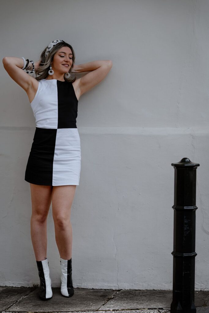 A woman poses against a plain wall, wearing a black-and-white colour-blocked dress, matching boots, and a patterned headscarf. She smiles with her arms raised, looking relaxed and confident. Copyright Ashley Kew Photography - All Rights Reserved