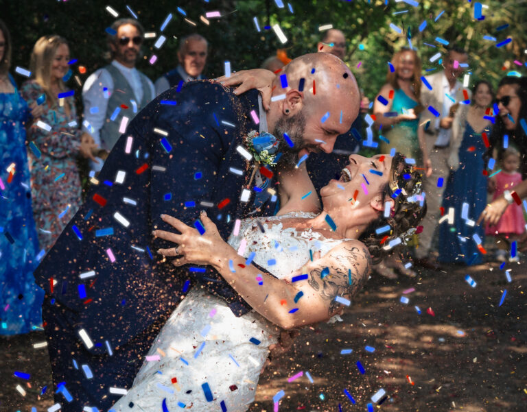 A joyful bride and groom share a dip and smile at each other as colourful confetti rains down. Guests in bright clothes cheer in the background during an outdoor wedding celebration. Copyright Ashley Kew Photography - All Rights Reserved