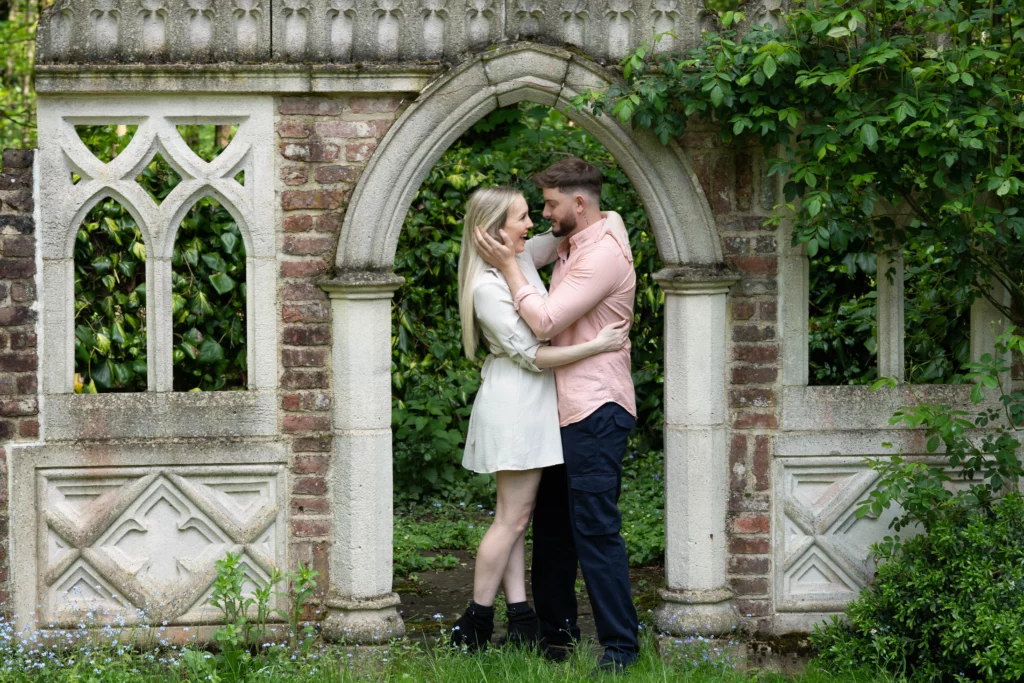 A couple stands closely facing each other, smiling and embracing under a stone archway with gothic-style windows, surrounded by greenery and plants. Copyright Ashley Kew Photography - All Rights Reserved