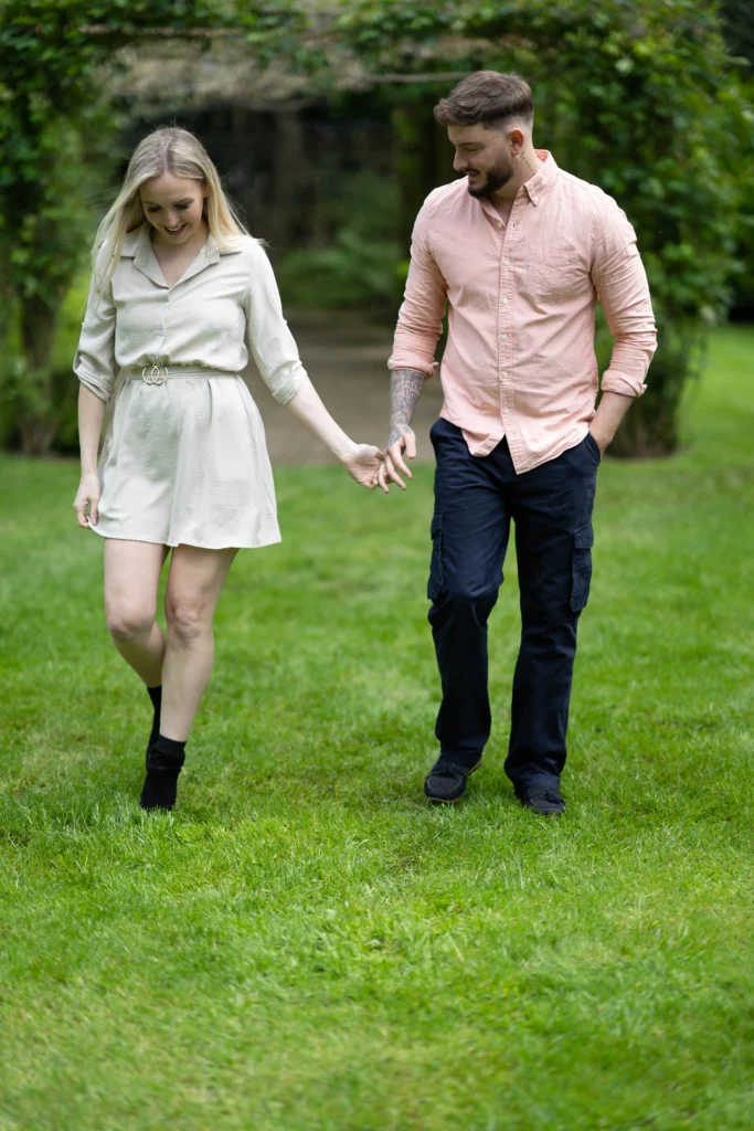 A smiling couple walk hand-in-hand on green grass. The woman wears a light dress and black boots, while the man wears a peach shirt and dark trousers. Trees and greenery are visible in the background. Copyright Ashley Kew Photography - All Rights Reserved